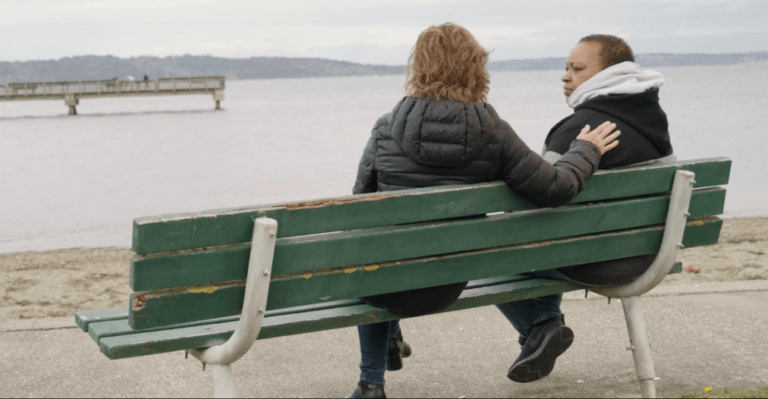 Two people sitting on a green bench overlooking a body of water